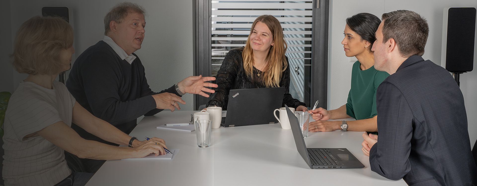 Five colleagues in a client meeting around a conference table with laptops